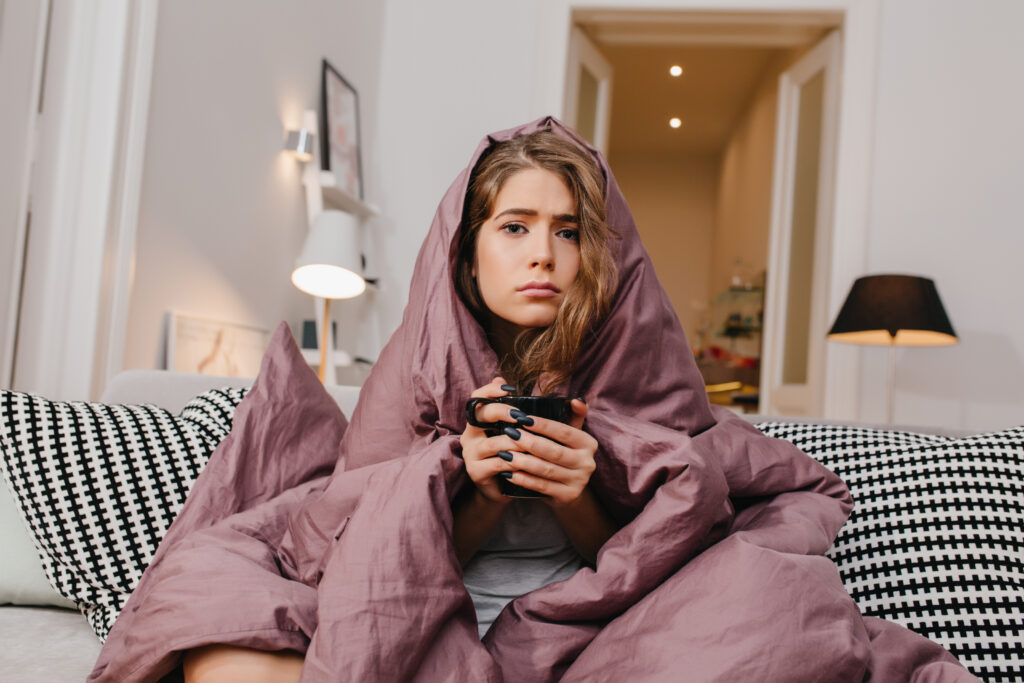 A young woman sits on a couch wrapped in a mauve blanket, looking unwell and holding a mug with both hands. She has a sad expression and is surrounded by soft home lighting.