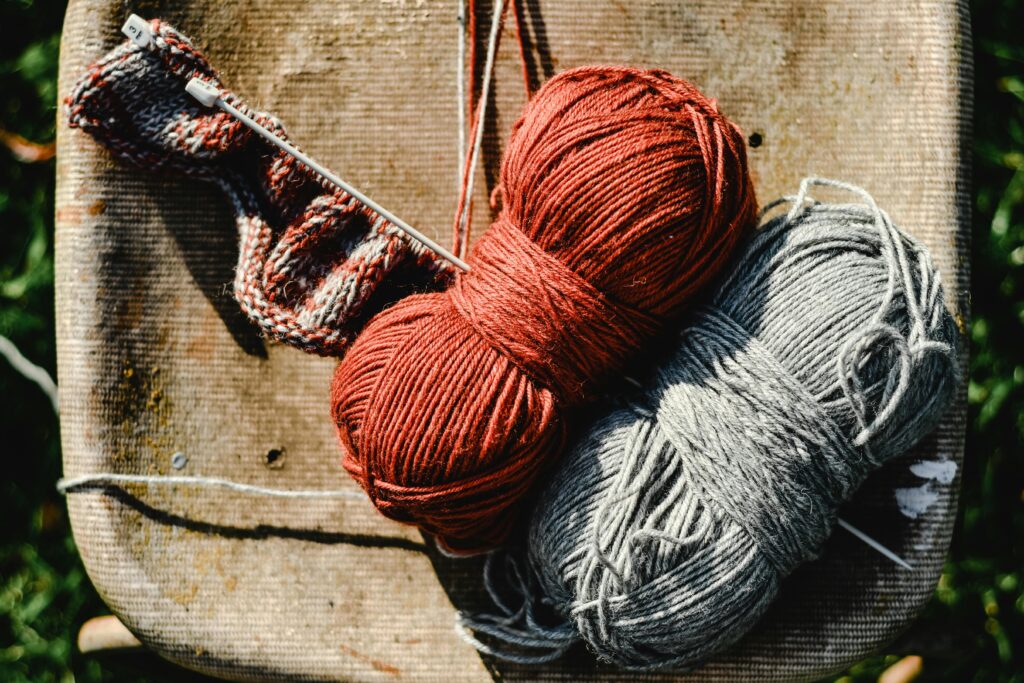 Two balls of yarn, one red and one gray, sit on a rustic stool next to a metal knitting needle with a partially knitted piece. The setting appears outdoors, with sunlight and green grass visible in the background.