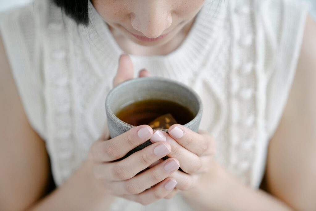 A woman wearing a white knit sweater holds a gray ceramic cup of tea with both hands, close to her face, as she appears to enjoy the warmth and aroma. Her nails are neatly manicured with pale pink polish.