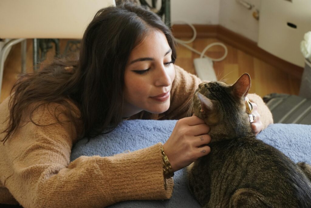 A woman with long brown hair lies on a bed, gently petting a tabby cat and looking at it affectionately. The room has wooden floors and a cozy, relaxed atmosphere.