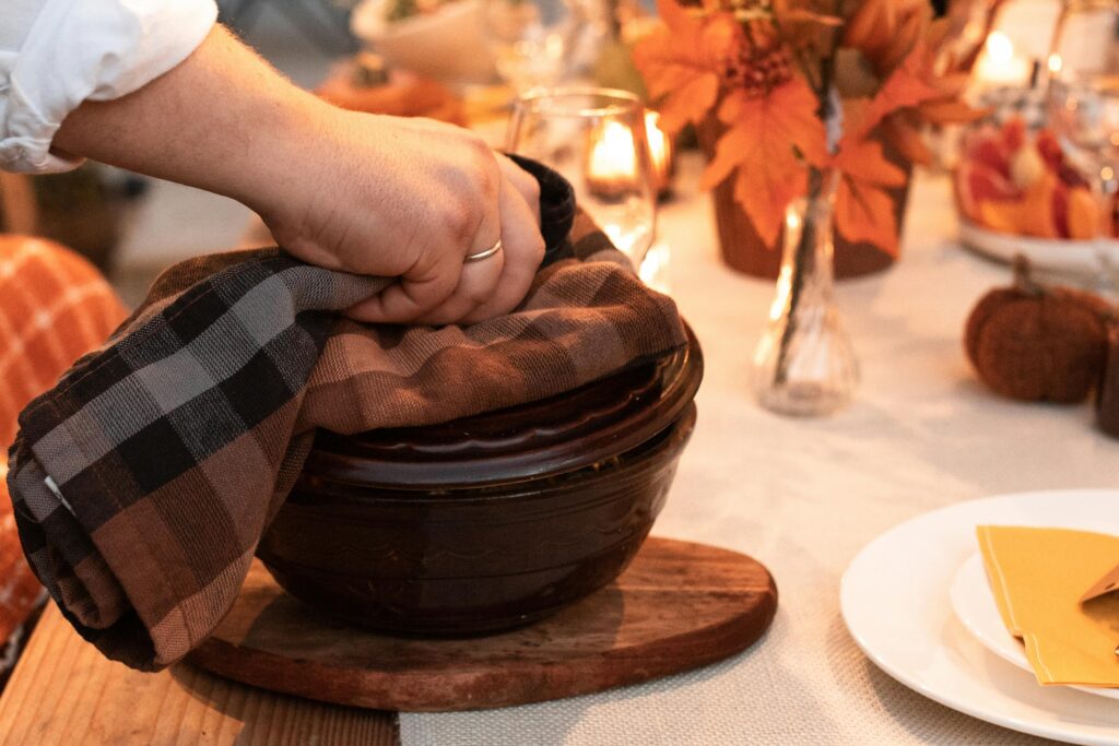 A hand with a gold ring lifts a plaid cloth from a ceramic bowl on a wooden board, set on a table decorated with fall leaves, candles, and pumpkins.