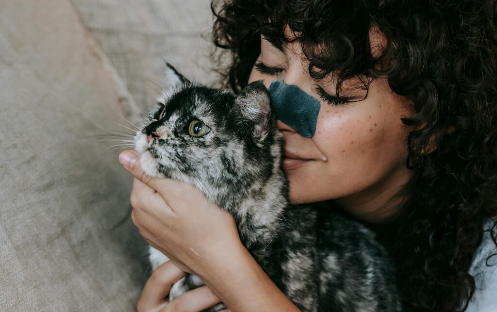Woman with curly hair affectionately cuddling a gray tortoiseshell cat, with a blue nose strip on her face and a gentle smile.