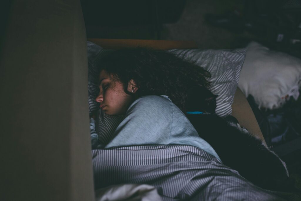 A person with curly hair sleeps on a bed, facing left, under a striped blanket. A black cat lies close behind them, also resting. The lighting is dim and the setting appears calm and cozy.